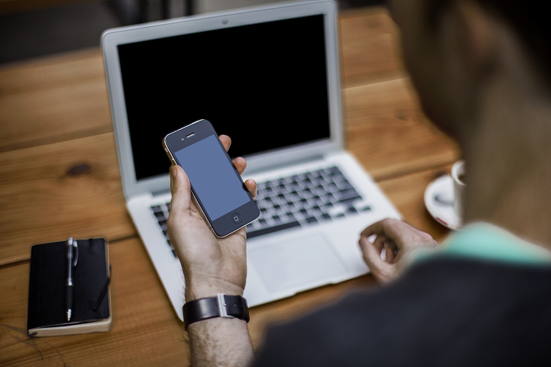 personne qui consulte son smartphone devant un ordinateur portable ouvert, carnet et stylo posés sur le bureau
